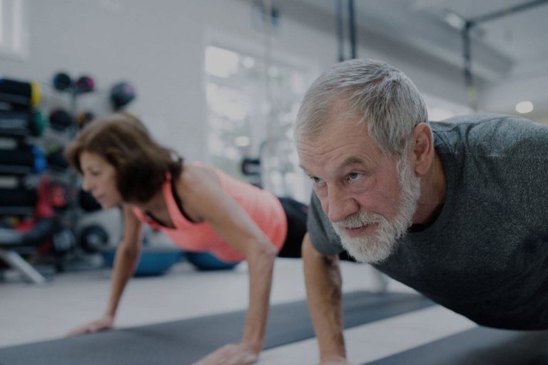Billede af to ældre personer der udfører nogle træningsøvelser på en yogamåtte.